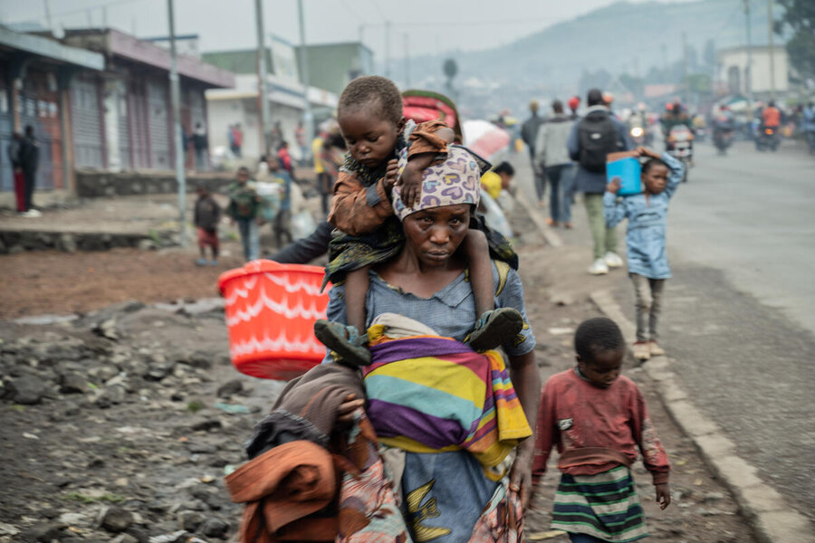 A displaced woman carries her child through rubble-strewn streets, belongings wrapped in cloth, as families flee conflict in eastern DR Congo