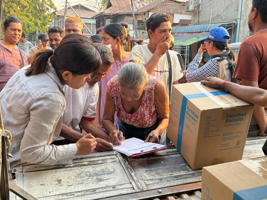 A woman signs a document on a clipboard at a food distribution point, with a WFP box of food assistance in view