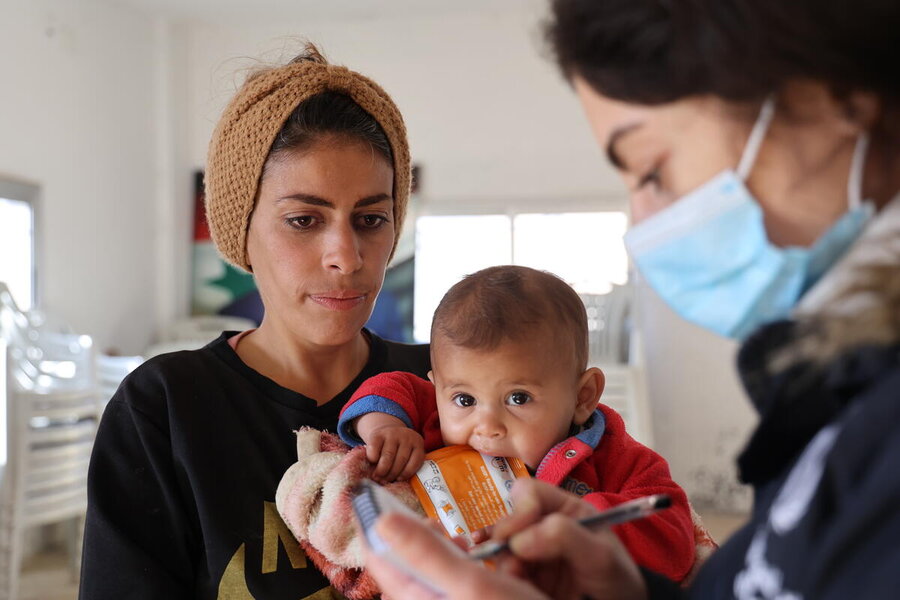 A baby looks into camera, held by her mother, as  nutritionist looks on wearing a facemask