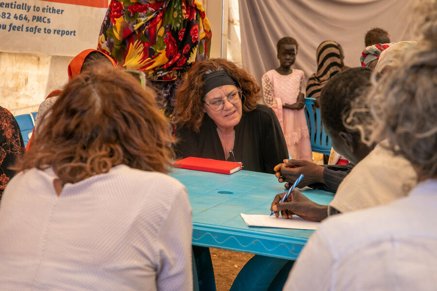 A white woman at a blue plastic table in a tend talks to women in Renk, South Sudan