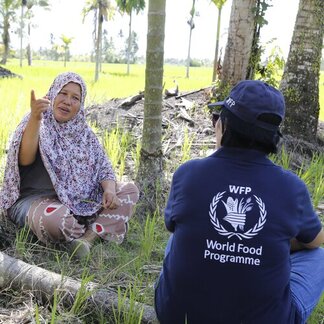 Person in headscarf points upwards while sitting in a field. Another person in a WFP shirt sits opposite. Green fields and trees are in the background.