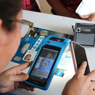 Person scans QR code on smartphone using blue device. Table holds another device and printed materials.