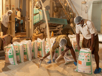 A worker bent over scooping grain while another holds a sack, in a rice fortification mill in Gajapati